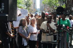 Kizito Musabimana standing beside Debbie Hill-Corrigan and Loly Rico speaking to cameras with a crown behind them at 129 Peter Street