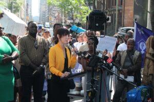 Diane Walter standing behind a podium speaking to cameras with a crown behind her at 129 Peter Street