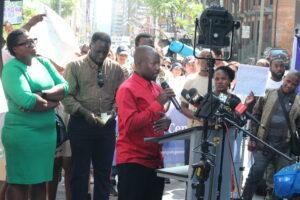 Mr. Asuman standing behind a podium speaking to cameras with a crown behind him at 129 Peter Street