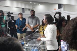 Pastor Judith speaking behind a podium to cameras with a crowd behind her in Revivaltimes Tabernacle Church