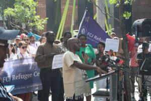 Kizito Musabimana standing behind a podium speaking to cameras with a crown behind him at 129 Peter Street