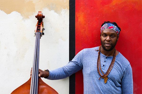 Jonathan Michel smiles and holds his upright bass against a red and white wall.