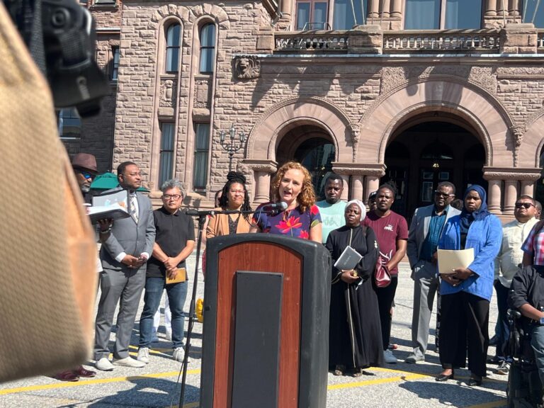 Rev. Alexa Gilmour standing at a podium, addressing a crowd of people at Queen's Park, with the park's iconic architecture visible in the background.